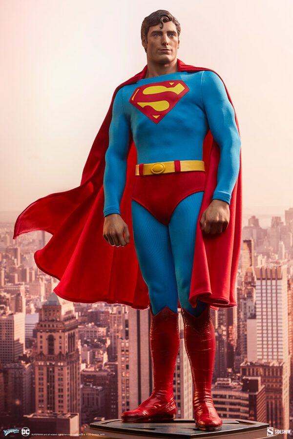 Superman: The Movie Premium Format™ Figure from Sideshow Collectibles and DC Comics. A statue of an iconic superhero with a red cape and blue costume stands overlooking a cityscape background.