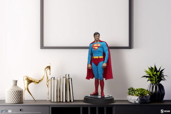 Superman: The Movie Premium Format™ Figure from Sideshow Collectibles and DC Comics. A Superman figurine standing on a sleek shelf, with framed art above, books, a golden sculpture, and potted plants nearby.