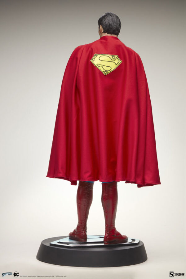 Superman: The Movie Premium Format™ Figure from Sideshow Collectibles and DC Comics. A collectible statue of a superhero, viewed from behind, showcasing a red cape with iconic yellow and red emblem, atop a display stand.