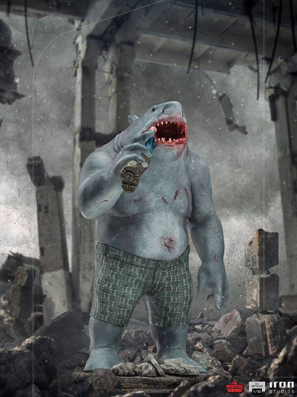 King Shark 1:10 Scale Statue from Iron Studios and DC Comics. Digital artwork of an anthropomorphic shark character dressed in shorts standing amidst rubble, with a menacing expression and a grenade in hand.