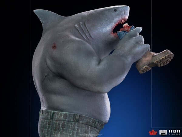 King Shark 1:10 Scale Statue from Iron Studios and DC Comics. A 3D-rendered image of an anthropomorphic shark character standing upright and eating a human hand, dressed in plaid pants and shoes, with a dark background.