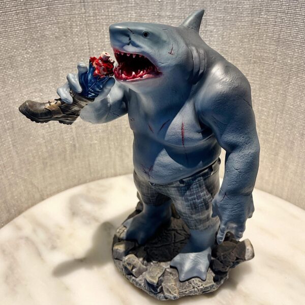 King Shark 1:10 Scale Statue from Iron Studios and DC Comics. A collectible figurine of an anthropomorphic shark creature standing upright, wearing shorts and biting into a severed fake leg.