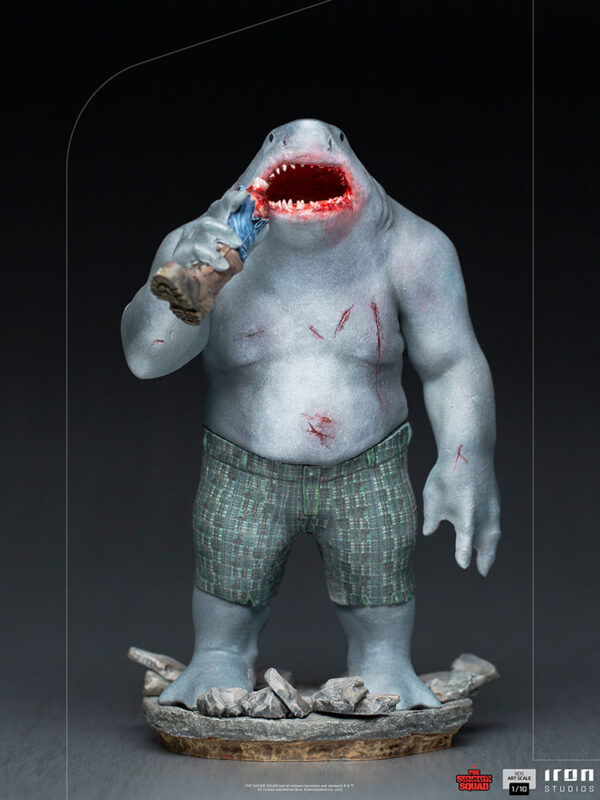 King Shark 1:10 Scale Statue from Iron Studios and DC Comics. Collectible figurine of a humanoid with a shark head, wearing shorts, standing on a rocky base, and biting into a small figure.