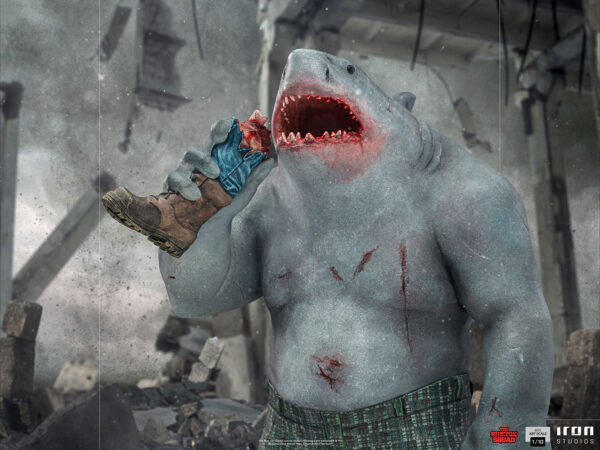 King Shark 1:10 Scale Statue from Iron Studios and DC Comics. Illustration of a shark-headed humanoid creature with a mutilated limb in its mouth amidst a destroyed environment, showcasing a chaotic and menacing scene.