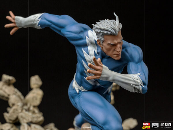 Marvel Quicksilver 1:10 scale statue from Iron Studios. An action figure of a superhero in a dynamic pose with white hair and a blue suit, set against a black background with blurred rocks at the base.