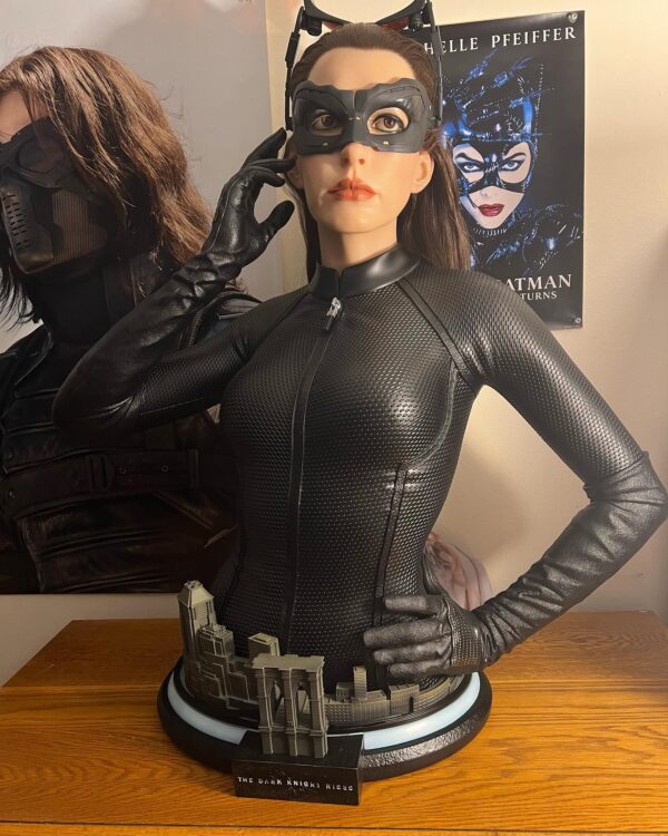 Catwoman (Selina Kyle) Life-Size Bust by Infinity Studio X Penguin Toys. Life-size bust of Catwoman (Selina Kyle), as portrayed by Anne Hathaway, wearing a sleek, textured black catsuit with her signature mask and cat ears. She stands in a poised stance with one hand adjusting her mask, capturing a determined expression. The base features miniature Gotham City buildings, including an archway labeled 'The Dark Knight Rises.'