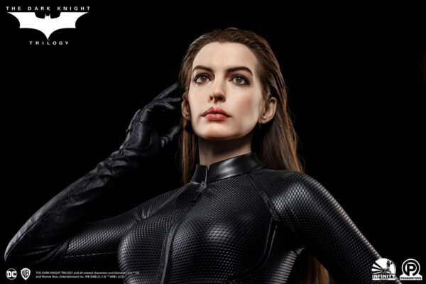 Catwoman (Selina Kyle) Life-Size Bust by Infinity Studio X Penguin Toys. Life-size bust of Catwoman (Selina Kyle), as portrayed by Anne Hathaway, wearing a sleek, textured black catsuit with her signature mask and cat ears. She stands in a poised stance with one hand adjusting her mask, capturing a determined expression. The base features miniature Gotham City buildings, including an archway labeled 'The Dark Knight Rises.'