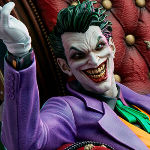 The Joker Deluxe Collector Edition Maquette from DC Comics and Tweeterhead. Close-up of a detailed Joker figure with a wide, menacing smile, wearing a purple suit and sitting in front of a red cushioned background.