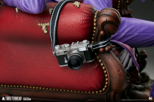 The Joker Deluxe Collector Edition Maquette from DC Comics and Tweeterhead. A detailed view of a miniature camera with a striped strap resting on a cushioned vintage chair, partially obscured by a purple fabric suggestive of the Joker character from DC Comics.