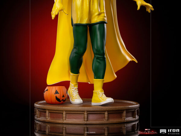 Vision Halloween Version WandaVision Art Scale 1:10 Statue from Marvel and Iron Studios. A collectible figure of a superhero in a yellow cape and green suit standing beside a Halloween pumpkin on a wooden stand.